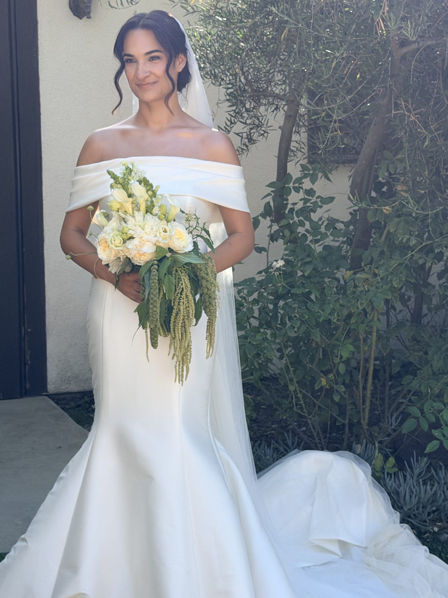 Bride standing with bouquet, smiling by olive tree
